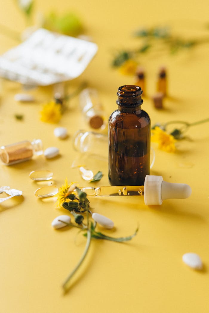 about-01 Brown glass dropper bottle among herbs and pills on yellow surface, ideal for wellness themes.